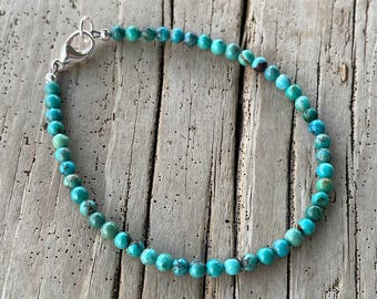 May include: A turquoise beaded bracelet with a silver clasp. The bracelet is made of small, round, turquoise beads. The beads have variations in color, with some showing darker blue-green hues. The bracelet is displayed on a wooden surface.