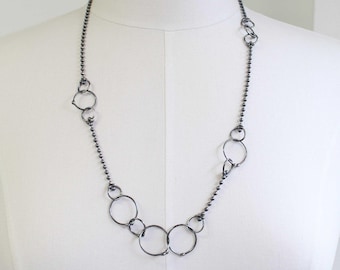 May include: A long, dark gray necklace featuring a series of interconnected open circles and small ball chain links. The necklace is displayed on a white mannequin, showcasing its design and length. The circles vary in size, creating a unique, modern aesthetic.