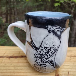 May include: White ceramic mug with a blue jay illustration. The mug has a white handle and a light blue interior. The rim is black. The mug is sitting on a wooden surface.