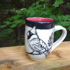 May include: White ceramic mug with a black rim and handle, featuring a black and white illustration of a cardinal bird. The interior of the mug is red. The mug is sitting on a wooden surface.