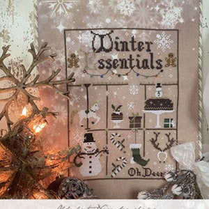 May include: A cross-stitch pattern featuring a winter-themed design with a snowman, reindeer, and other holiday elements. The pattern is titled "Winter Essentials" and is part of the 2023 Nashville Collection by The Primitive Hare.