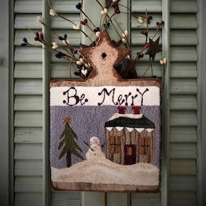 May include: A rustic, rectangular Christmas decoration with the words "Be Merry" stitched across the top. The design features a house, snowman, and Christmas tree in a snowy landscape. The decoration is adorned with star-shaped accents and berry branches.