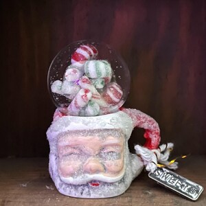 May include: A snow globe featuring a Santa Claus base with a clear globe filled with red and white striped candies. The base is white with glitter, and a small tag that says "Sweets" hangs from the side.
