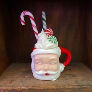 May include: A ceramic Santa mug with a red handle and white whipped cream topping. Two candy canes and two candies are placed in the whipped cream. The mug is white with Santa's face and a red rim.