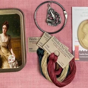 May include: A collection of vintage-style needlework supplies on a pink linen surface. Includes a tin with a painting, a silver charm, embroidery floss, and a cameo. The Weeks Dye Works floss is in various colors.