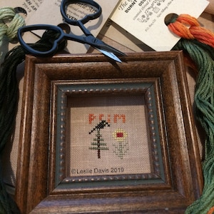 May include: A framed cross-stitch design with a small tree and a bird, with the text "PRIM" above the tree and "Leslie Davis 2019" below. The frame is brown wood with a decorative border.