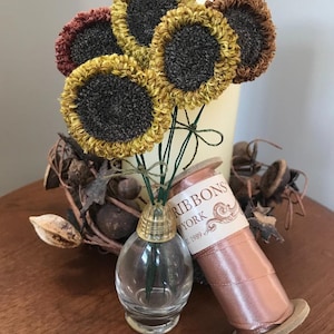May include: A bouquet of five small, handmade felt sunflowers in shades of brown, yellow, and orange. The sunflowers are arranged in a clear glass vase with a gold top. A spool of pink ribbon with the text "Ribbons New York Est. 1989" is on a wooden surface.
