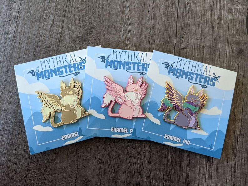 May include: Three enamel pins featuring mythical creatures with wings. Each pin has a different colour scheme and creature design. The pins are on a blue background with white clouds and the text "Mythikal Monsters" and "Enamel Pin".
