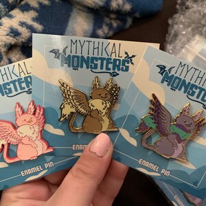 May include: Three enamel pins featuring mythical creatures with wings. The pins are on blue and white cards with the text "Mythikal Monsters" and "Enamel Pin". The creatures are a pink cat-like creature, a brown cat-like creature, and a purple cat-like creature.