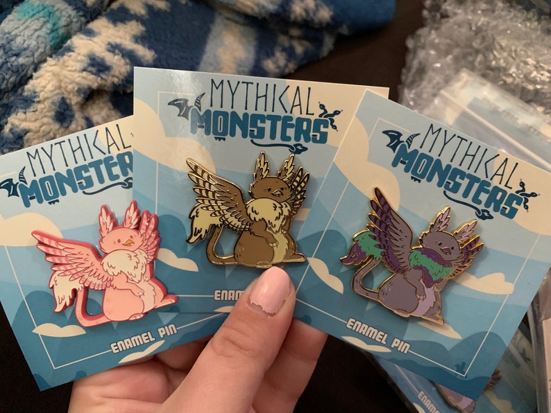 May include: Three enamel pins featuring mythical creatures with wings. The pins are on blue and white cards with the text "Mythikal Monsters" and "Enamel Pin". The creatures are a pink cat-like creature, a brown cat-like creature, and a purple cat-like creature.