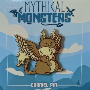 May include: Gold enamel pin depicting a mythical creature with brown fur, yellow wings, and a beak. The creature is sitting on its haunches with its wings spread out. The pin is displayed against a blue and white background with the text "Mythikal Monsters" and "Enamel Pin".
