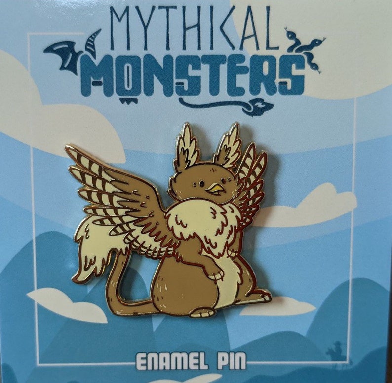 May include: Gold enamel pin depicting a mythical creature with brown fur, yellow wings, and a beak. The creature is sitting on its haunches with its wings spread out. The pin is displayed against a blue and white background with the text "Mythikal Monsters" and "Enamel Pin".
