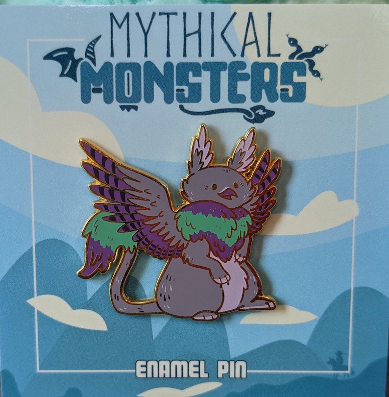 May include: A purple and green enamel pin of a mythical creature with wings. The creature has a grey body, a purple and green feathered mane, and purple and gold wings. The pin is on a blue and white background with the text "Mythikal Monsters" and "Enamel Pin".