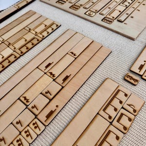 May include: A light-coloured wooden rhythm composition board set featuring laser-engraved musical notes, rests, and rhythmic symbols. The set includes a main board labelled Rhythm Composition Board and various rectangular tiles with musical notation.