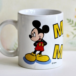 May include: White ceramic mug featuring Mickey Mouse in a classic pose. Mickey is wearing red shorts, yellow shoes, and a black top. The mug has the word "M" in yellow. A perfect gift for Disney fans.
