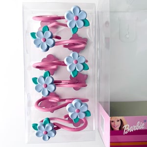 Può includere: Un set di fermagli per capelli rosa con decorazioni floreali blu e verdi. I fermagli sono in una confezione di plastica trasparente accanto a una scatola Barbie rosa e bianca. I petali dei fiori sono azzurri.