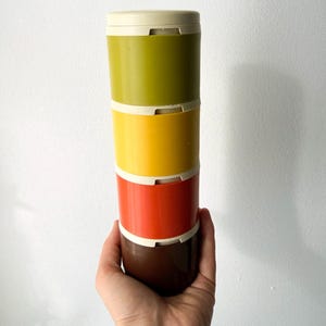 May include: A stack of four colorful, cylindrical containers in shades of olive green, yellow, orange, and brown, with off-white lids and connecting rings. The containers are held in a hand against a white background.