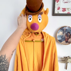 Pode incluir: Um macacão de fleece amarelo brilhante com um capuz que inclui um chapéu castanho, nariz roxo e olhos brancos grandes. A peça tem um fecho de correr frontal, mangas compridas com punhos brancos e detalhes pretos em forma de garras.