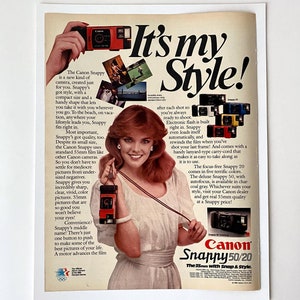 Peut inclure: Publicité pour l'appareil photo Canon Snappy 50/20. L'annonce présente une femme tenant un appareil photo rouge. Le texte "It's my Style!" est affiché en grandes lettres en gras. D'autres appareils photo sont présentés en différentes couleurs.