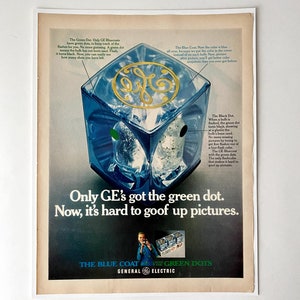 May include: A blue cube with a green dot on one side and a black dot on another side. The cube is a visual representation of a flashbulb. The text on the image reads "Only GE's got the green dot. Now, it's hard to goof up pictures." The text "The Blue Coat. The Green Dots. General Electric" is also visible.