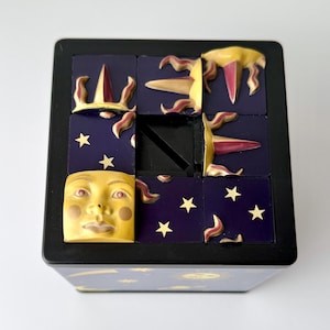 Könnte beinhalten: Ein dekorativer Puzzlewürfel mit schwarzem Rahmen. Die Würfelflächen zeigen ein gelbes Mondgesicht, Sterne und stilisierte Sonnen mit roten und gelben Akzenten auf dunkelblauem Hintergrund. Ein einzigartiges Objekt mit Himmelsmotiv.