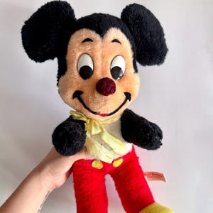 May include: Vintage Mickey Mouse plush doll with black ears, a peach-colored face, and a red nose. The doll wears red pants with yellow buttons and yellow feet. A yellow bow tie is around the neck. The doll is a classic Disney character.