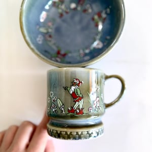 Puede incluir: Una taza de cerámica con asa y un cuenco a juego. La taza presenta una ilustración caprichosa de un gnomo en un entorno forestal con setas y flores. El cuenco tiene un estampado floral.
