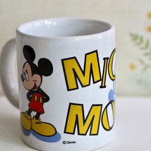 May include: White ceramic mug featuring Mickey Mouse. The mug has a handle and displays the name "Mickey Mouse" in yellow block letters. Mickey Mouse is depicted in his classic outfit, with red shorts and yellow shoes. The mug is a Disney product.
