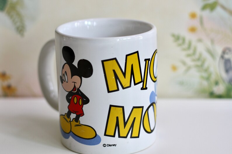May include: White ceramic mug featuring Mickey Mouse. The mug has a handle and displays the name "Mickey Mouse" in yellow block letters. Mickey Mouse is depicted in his classic outfit, with red shorts and yellow shoes. The mug is a Disney product.