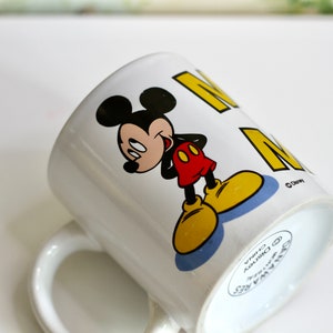 May include: White ceramic mug featuring Mickey Mouse in a classic pose, with black ears, a red shirt, yellow shoes, and a smiling expression. The mug has the word "M" in yellow. A Disney licensed product.
