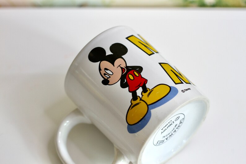 May include: White ceramic mug featuring Mickey Mouse in a classic pose, with black ears, a red shirt, yellow shoes, and a smiling expression. The mug has the word "M" in yellow. A Disney licensed product.