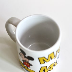 May include: White ceramic mug featuring a cartoon image of Mickey Mouse. The mug has a gray handle and the words "Mickey Mouse" in yellow and blue. A classic Disney-themed coffee mug.