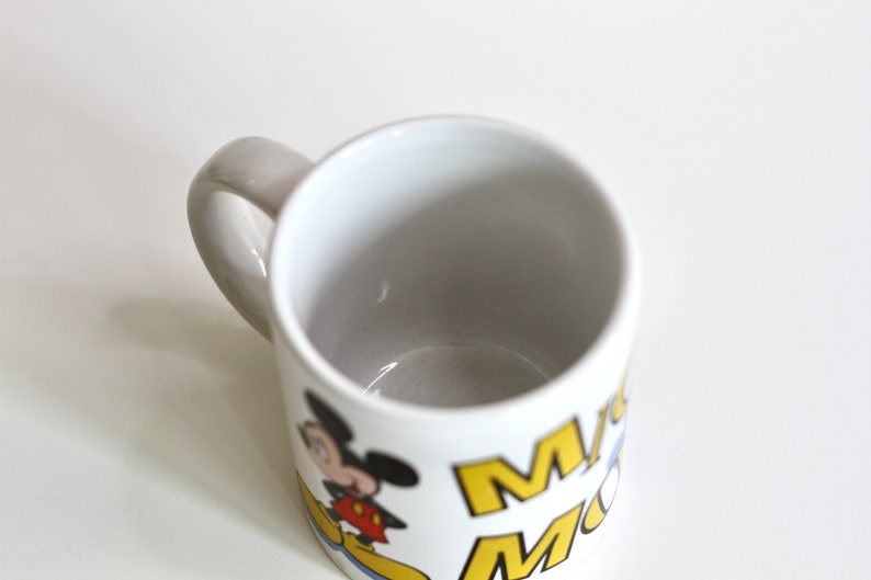 May include: White ceramic mug featuring a cartoon image of Mickey Mouse. The mug has a gray handle and the words "Mickey Mouse" in yellow and blue. A classic Disney-themed coffee mug.