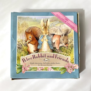 Peut inclure: Une boîte bleue avec une image de Peter Rabbit et deux écureuils. La boîte a un ruban rose avec le texte "200+ FULL-COLOR STICKERS" et le texte "Peter Rabbit and Friends Eight charming, full-color sticker books BY BEATRIX POTTER".