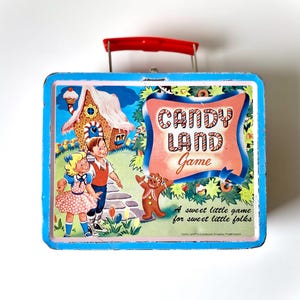 May include: A vintage blue and white tin game box with a red handle. The box features a colorful illustration of a gingerbread house, a gingerbread man, and two children. The text on the box reads "Candy Land Game" and "A sweet little game for sweet little folks".