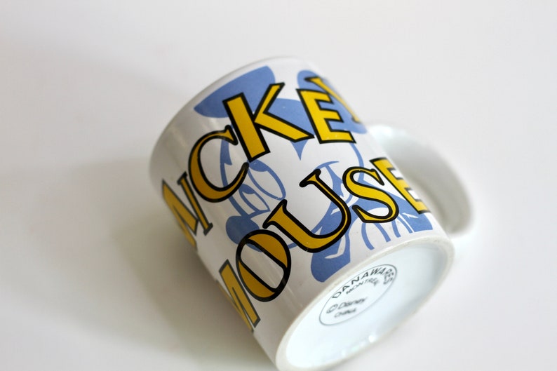 May include: White ceramic mug featuring the words "MICKEY MOUSE" in yellow outlined in black, with blue floral accents. The mug has a white handle and a Disney logo on the bottom. A classic Disney collectible.