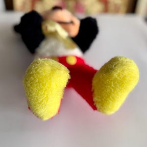 May include: A plush Mickey Mouse toy, lying on its back, with yellow feet, red shorts, and a white torso. The toy has black ears and a smiling face. A yellow button is visible on the shorts.