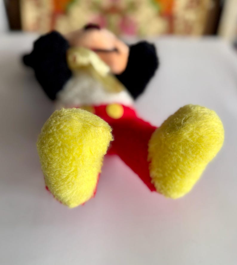 May include: A plush Mickey Mouse toy, lying on its back, with yellow feet, red shorts, and a white torso. The toy has black ears and a smiling face. A yellow button is visible on the shorts.