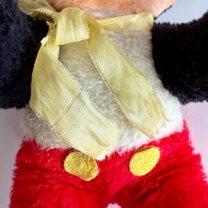 May include: Close-up of a vintage plush toy featuring a cartoon character. The toy has a peach-colored face, black ears and arms, and red pants with yellow buttons. A yellow ribbon is tied around the neck, and the body is white.