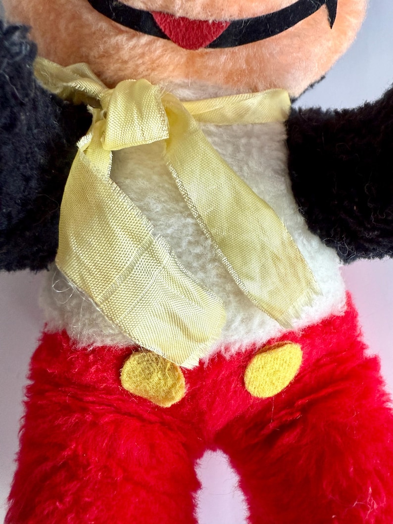 May include: Close-up of a vintage plush toy featuring a cartoon character. The toy has a peach-colored face, black ears and arms, and red pants with yellow buttons. A yellow ribbon is tied around the neck, and the body is white.