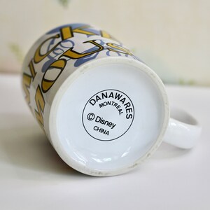May include: White ceramic mug with a handle, featuring a design with gold and blue lettering. The bottom of the mug has a circular logo with the text "DANAWARES MONTREAL &copy; Disney CHINA".