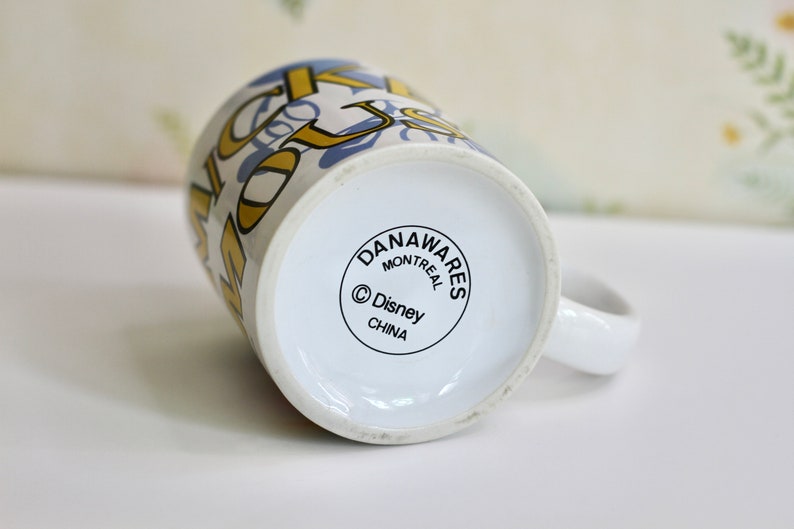 May include: White ceramic mug with a handle, featuring a design with gold and blue lettering. The bottom of the mug has a circular logo with the text "DANAWARES MONTREAL &copy; Disney CHINA".