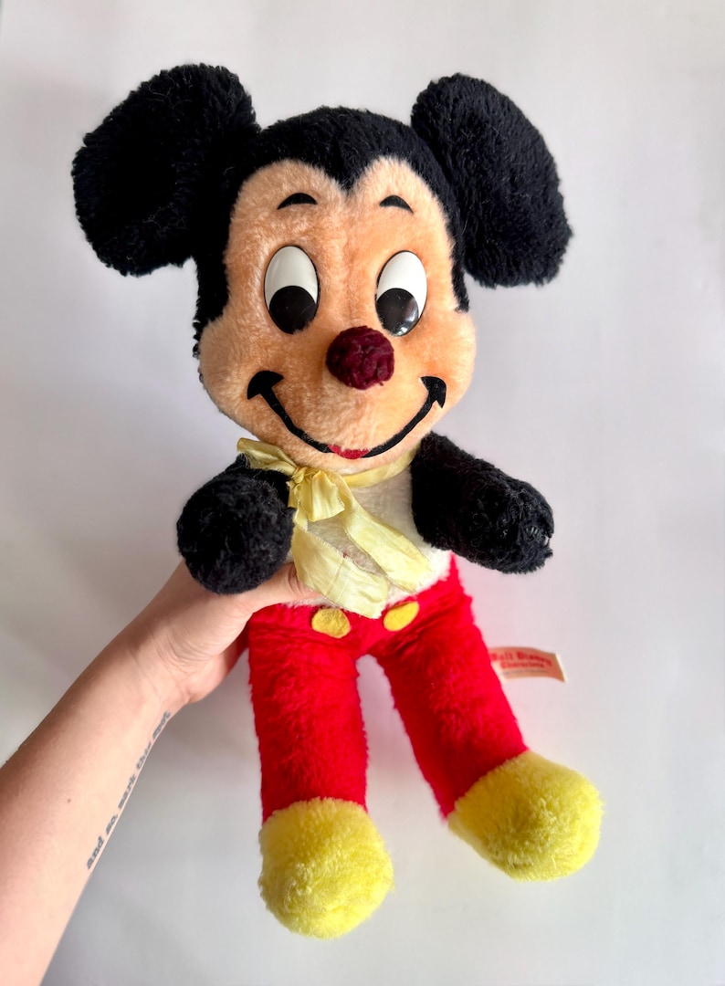 May include: Vintage Mickey Mouse plush doll with black ears, a peach-colored face, and a red nose. The doll wears red pants with yellow buttons and yellow feet. A yellow bow tie is around the neck. The doll is a classic Disney character.
