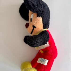 May include: A vintage Mickey Mouse plush toy. The plush features a black head and ears, a tan face, a red nose, and a red body. The toy has yellow feet and a yellow collar. A white tag is sewn on the red body.