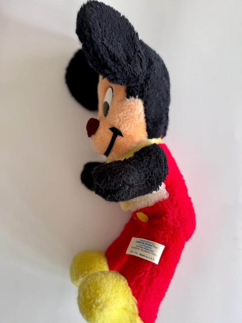 May include: A vintage Mickey Mouse plush toy. The plush features a black head and ears, a tan face, a red nose, and a red body. The toy has yellow feet and a yellow collar. A white tag is sewn on the red body.