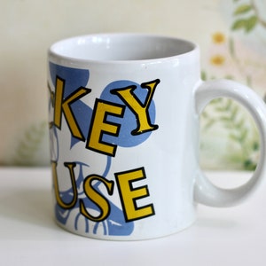 May include: White ceramic mug with the words "KEY MOUSE" in yellow and black lettering, set against a blue polka dot design. The mug has a curved handle and is likely for beverages.