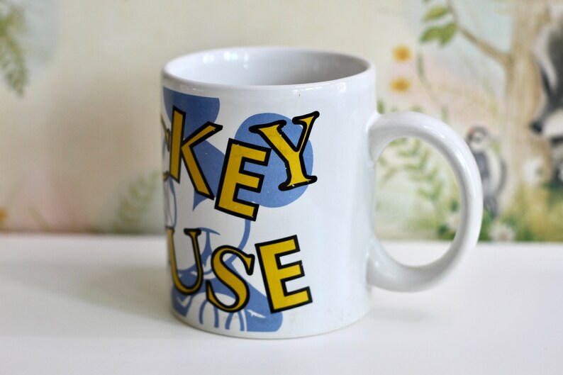 May include: White ceramic mug with the words "KEY MOUSE" in yellow and black lettering, set against a blue polka dot design. The mug has a curved handle and is likely for beverages.