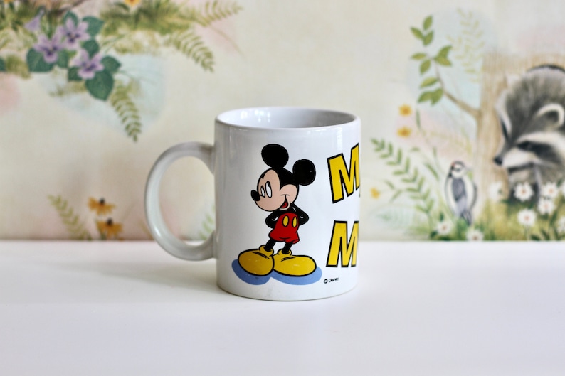 May include: White ceramic mug featuring Mickey Mouse in a classic pose. Mickey is wearing red shorts, yellow shoes, and a black top. The mug has the word "M" in yellow. A perfect gift for Disney fans.