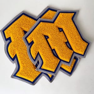 Könnte beinhalten: Drei gelbe Filzpatches mit blauem Rand, jeweils in Form des Buchstabens "M".