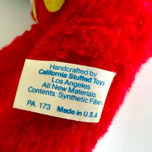 May include: Close-up of a red plush toy with yellow accents. A white tag is attached, displaying text: "Handcrafted by California Stuffed Toys, Los Angeles, All New Materials, Contents: Synthetic Fibers, PA. 173 Made in U.S.A."